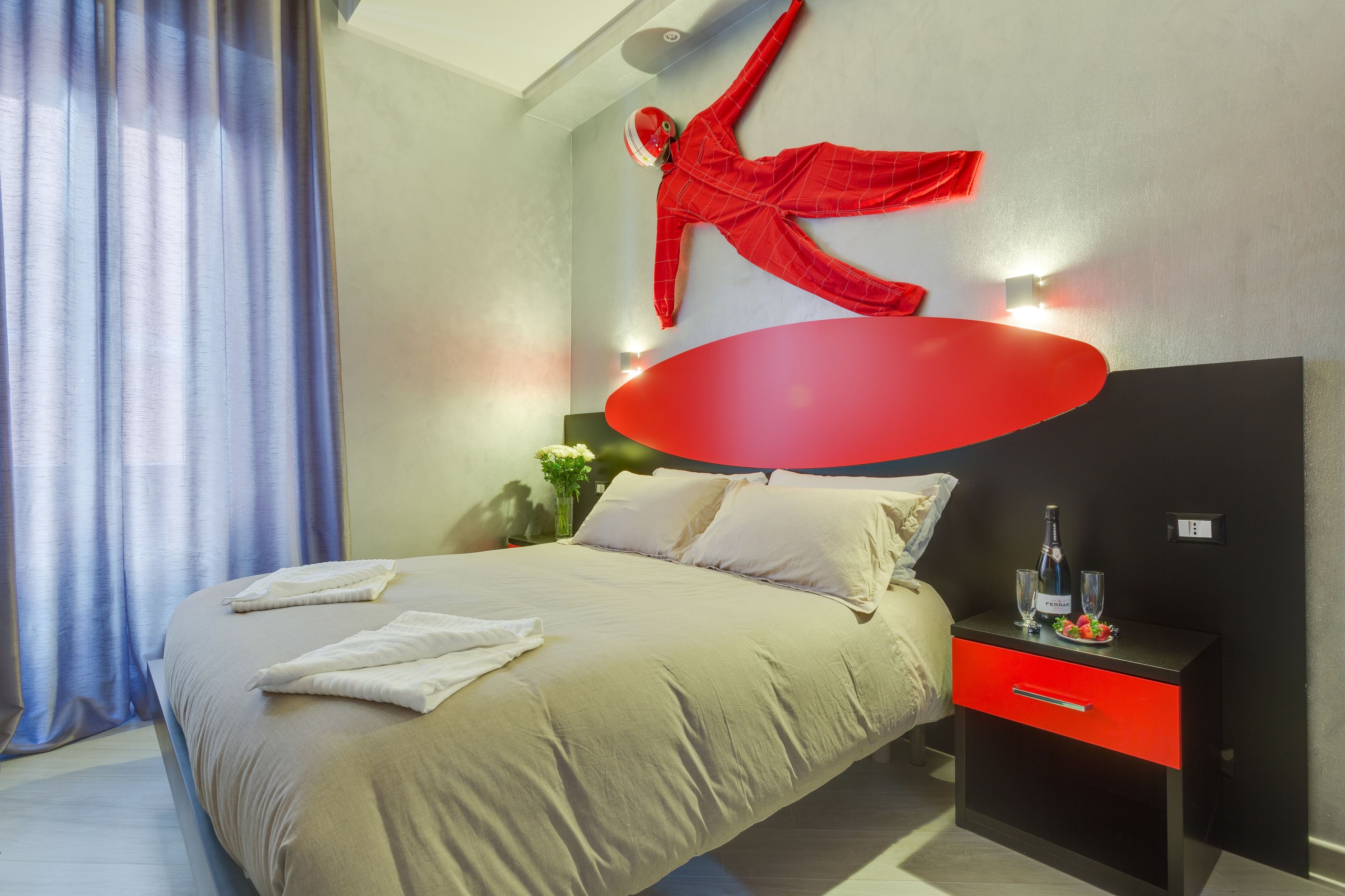 A modern hotel room with a large bed, a red accent wall, and a nightstand with a bottle and glasses.