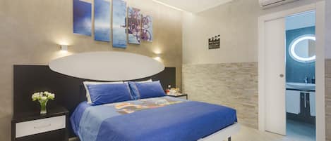 A modern bedroom with a large bed, bedside tables, a wall-mounted artwork, and a bathroom visible through an open door.
