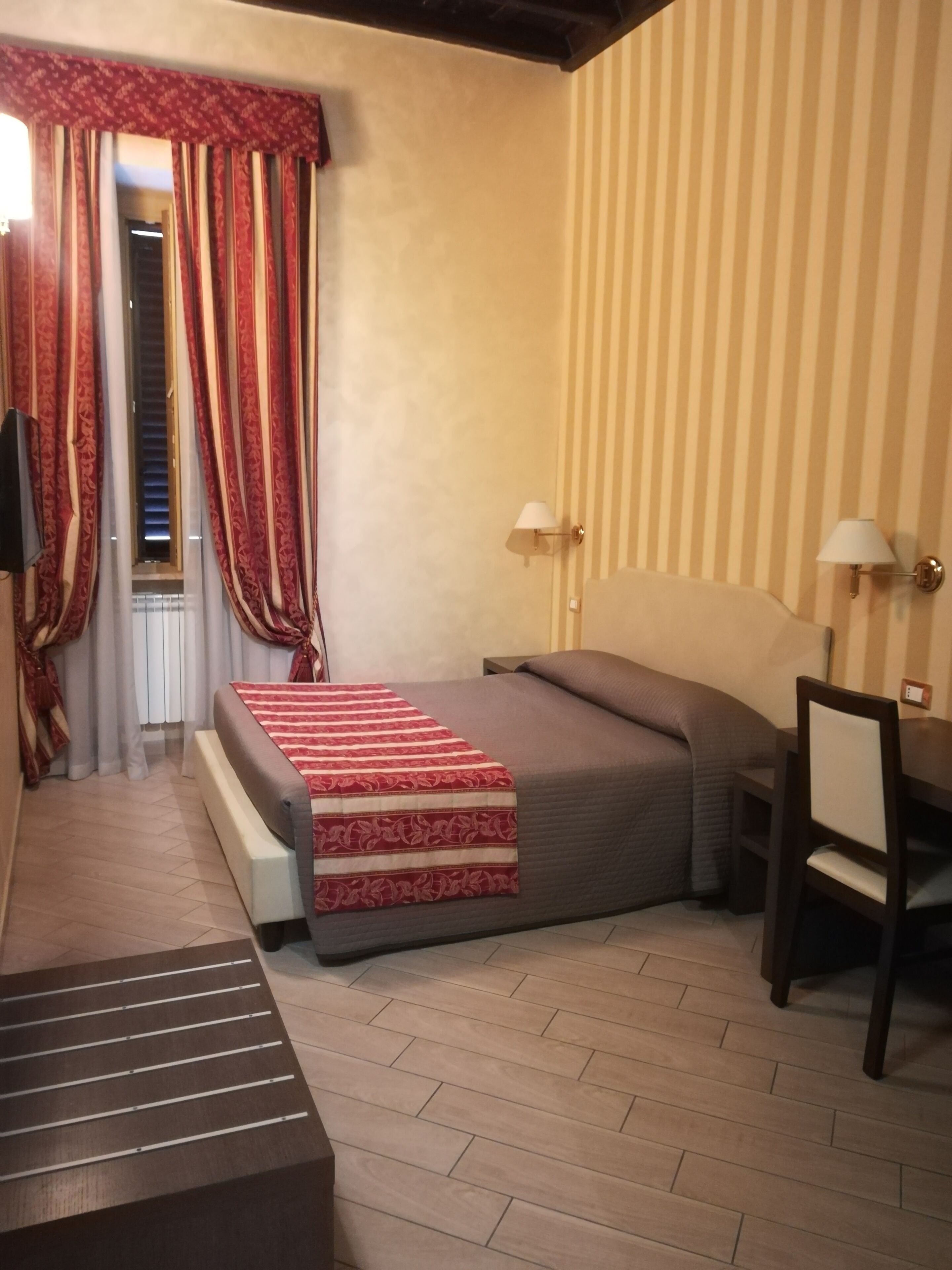 A hotel room with a bed, a desk, a chair, a lamp, and a window with curtains.