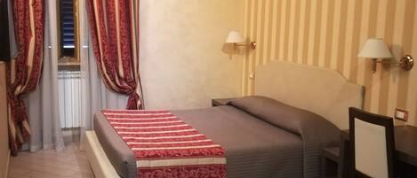A hotel room with a bed, a desk, a chair, a lamp, and a window with curtains.
