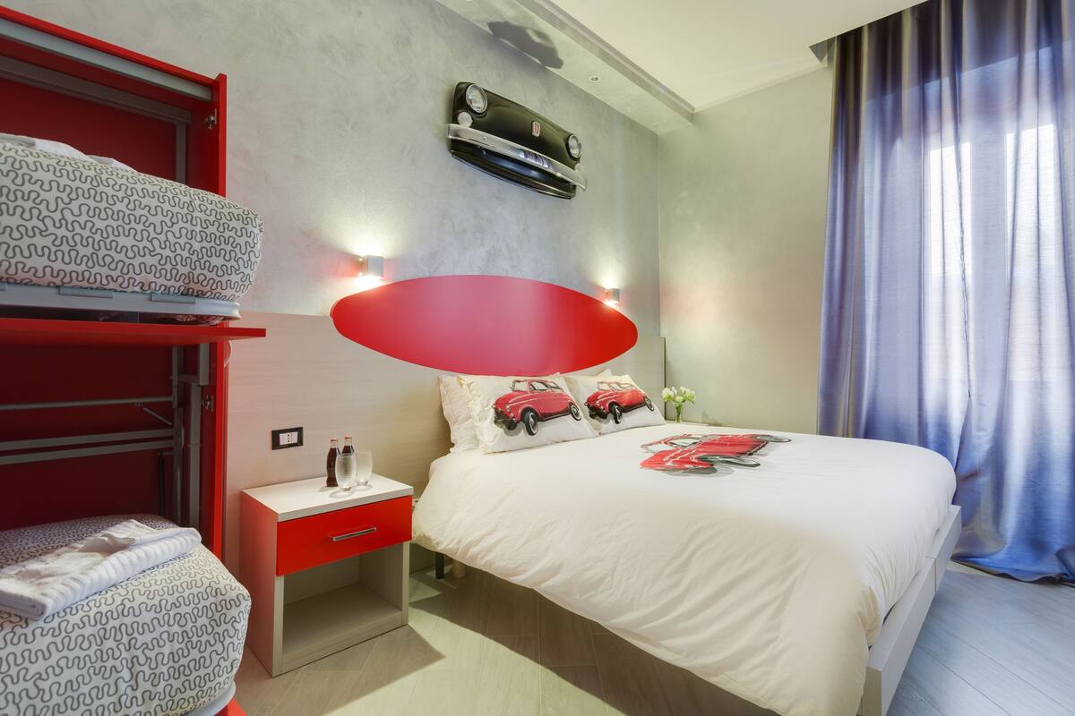 A hotel room with a bunk bed, a single bed with a red headboard, a bedside table with a lamp, and a window with curtains.