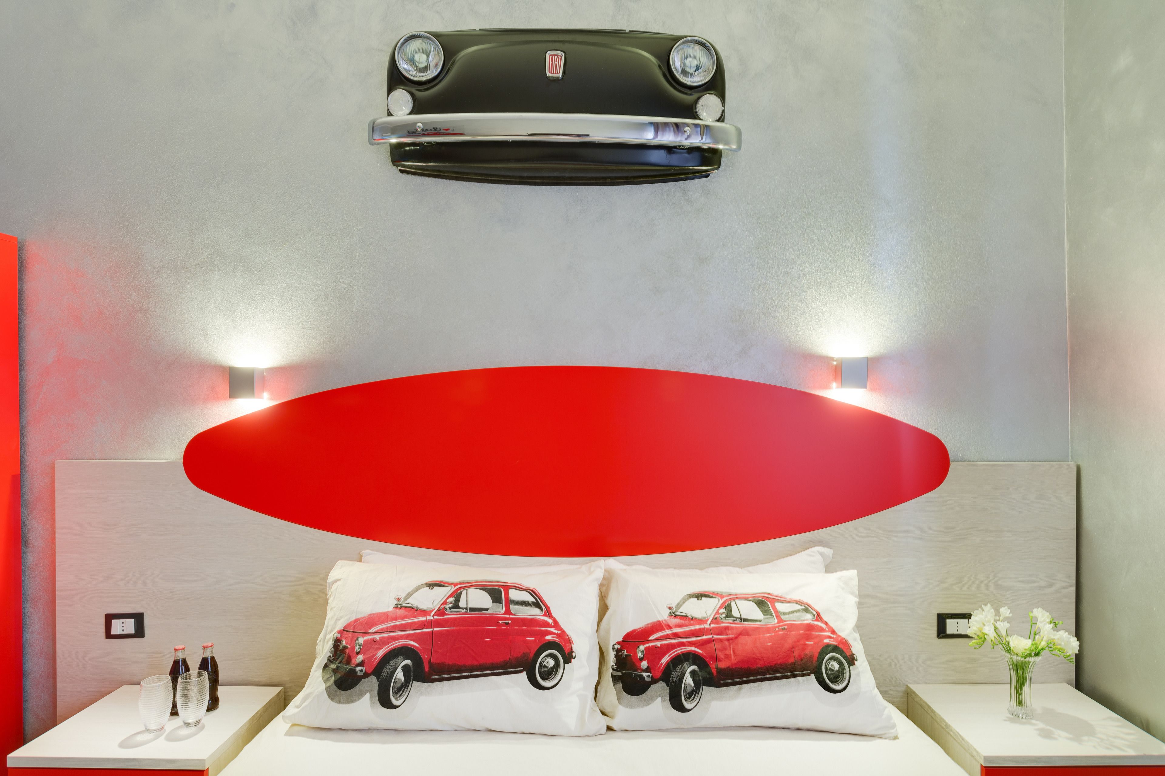 A hotel room with a modern design, featuring a large red oval light fixture above the bed, two pillows with a red car design, a small table with two bottles and glasses, and a wall-mounted air conditioning unit.
