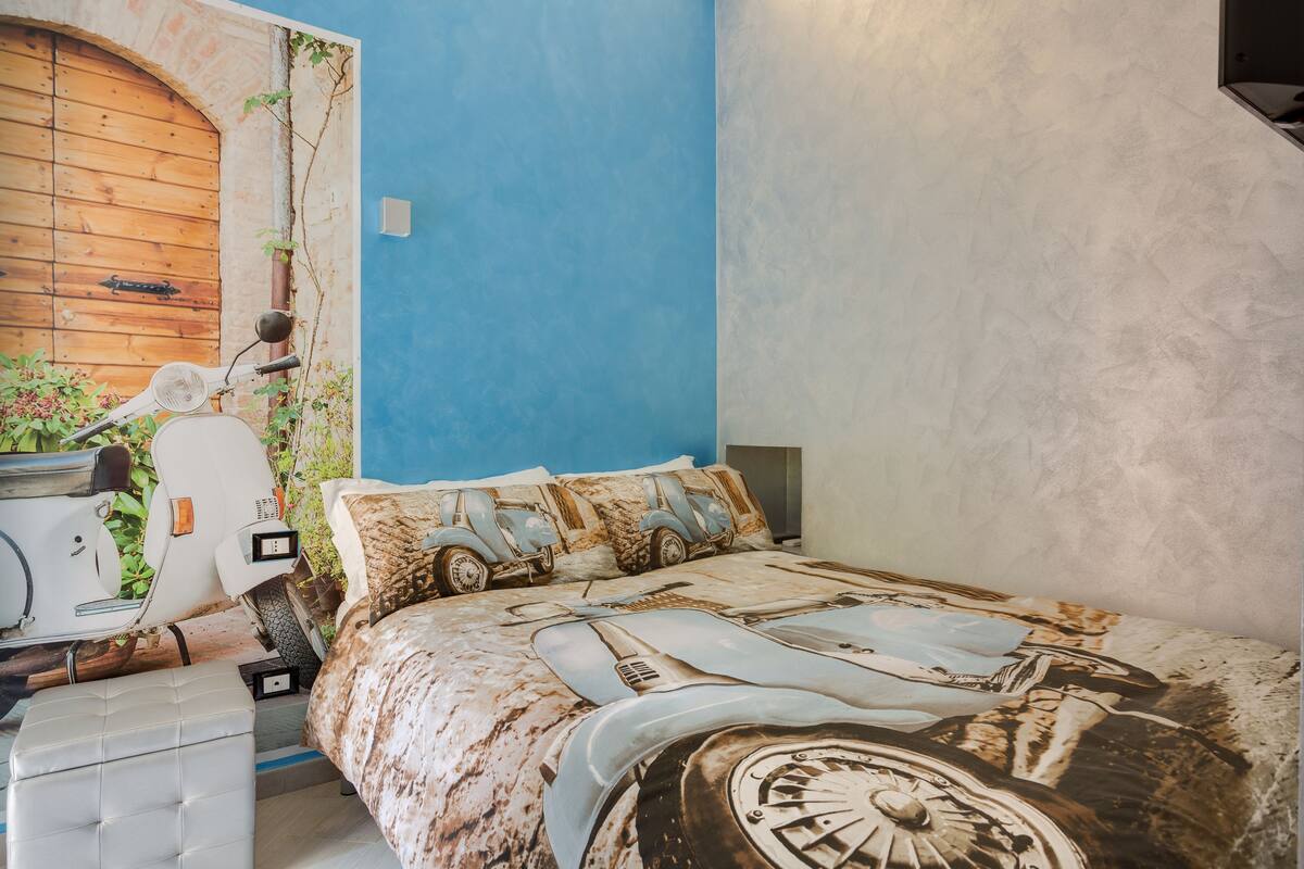 A bedroom with a blue wall, a bed with a vintage car design, a white scooter, and a wooden door.