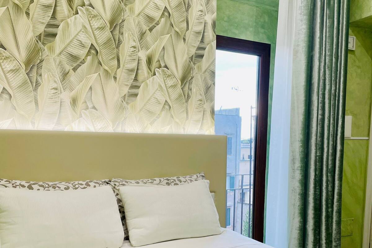 A neatly made bed with two pillows and two folded towels, a window with a view of buildings, and green curtains.