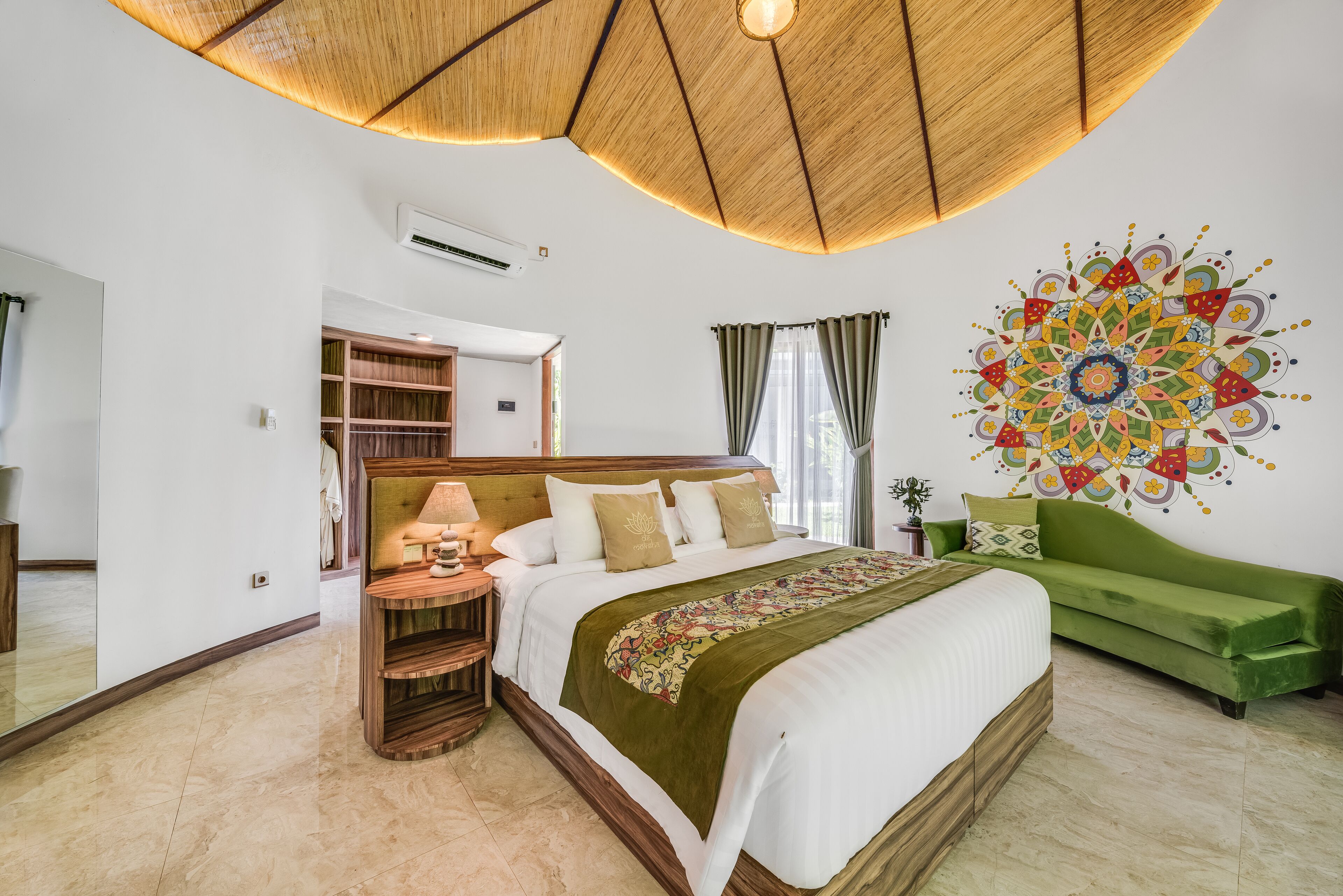 A bedroom with a large bed, a green sofa, a wooden headboard, a bedside table with a lamp, and a colorful wall decoration.