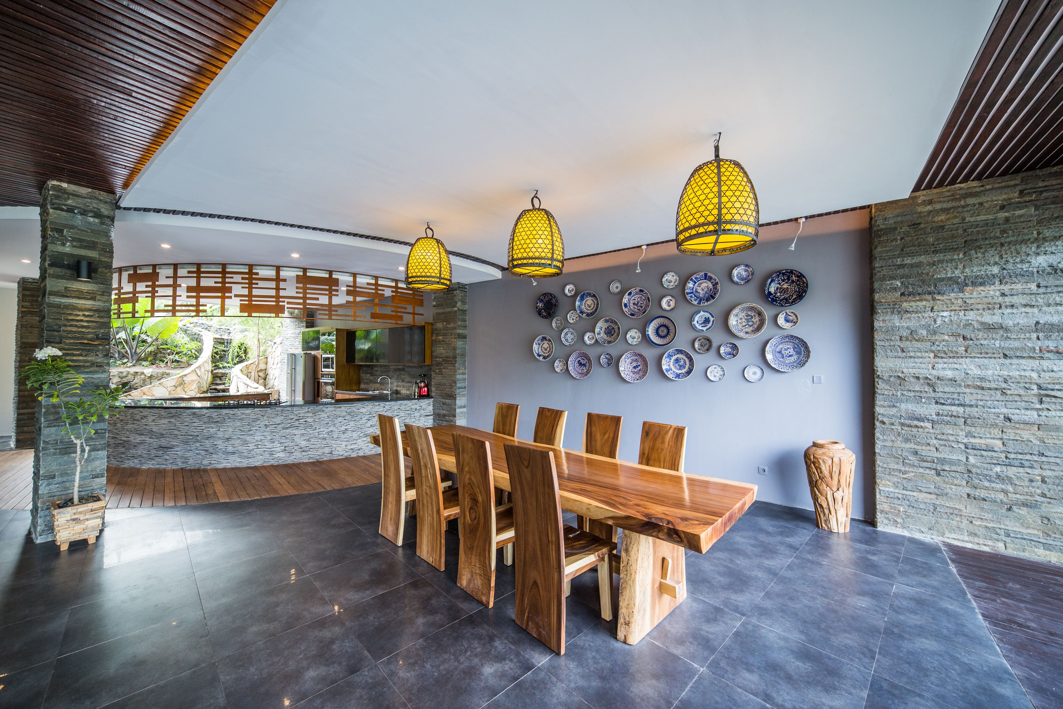 A modern dining area with a wooden table and chairs, a stone wall with decorative plates, and hanging yellow lamps.