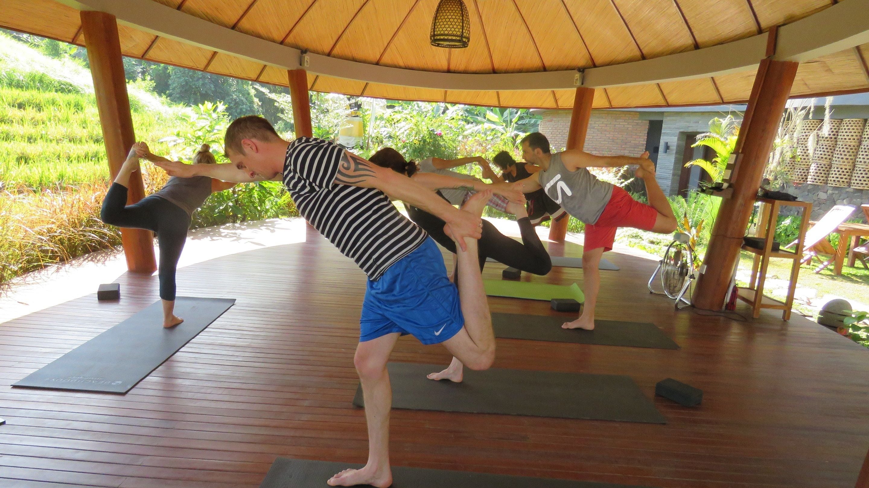 A group of people practicing yoga in an outdoor setting with wooden flooring and a thatched roof structure.