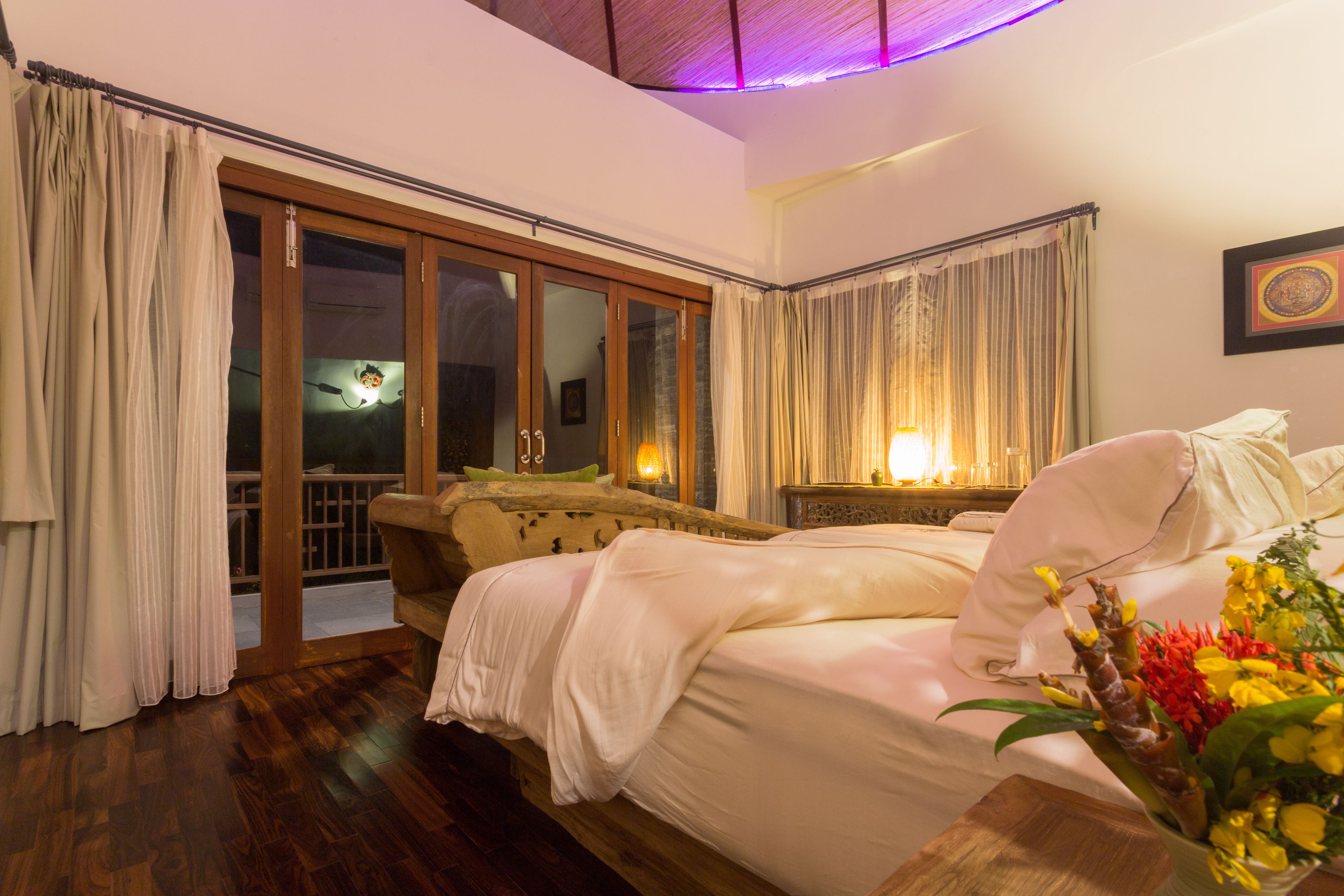 A bedroom with a wooden bed, a nightstand with a lamp, a window with curtains, and a view of the outside.