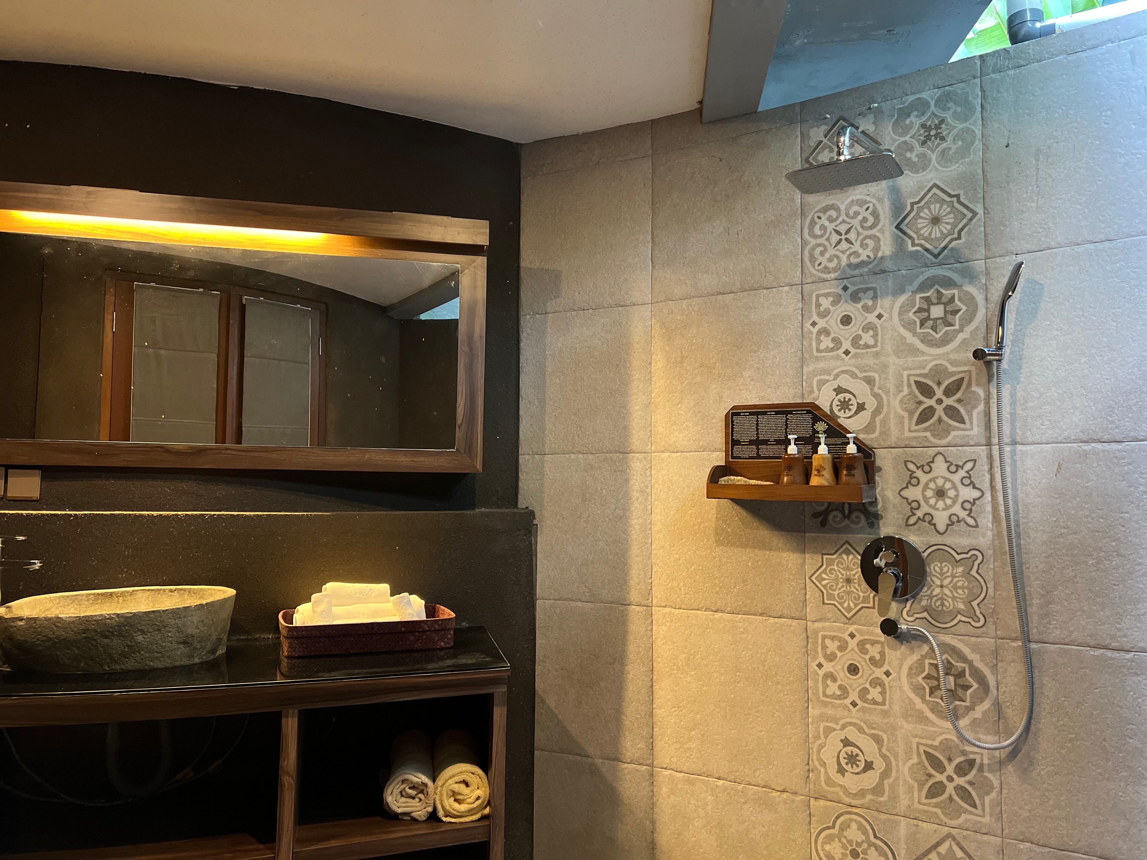 A bathroom with a large mirror, a wooden shelf with toiletries, a stone basin, and a shower with patterned tiles.