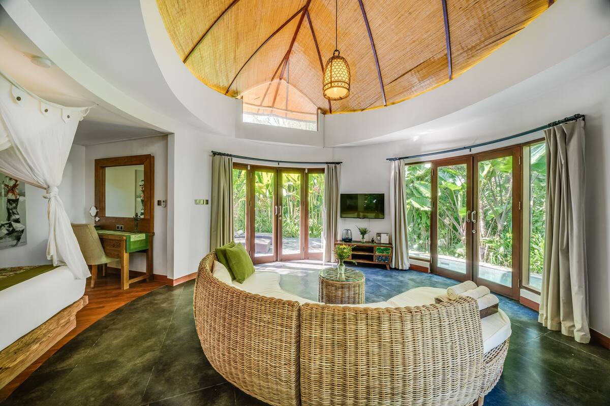 Ein geräumiger Wohnbereich mit einem gebogenen Rattan-Sofa, einem kleinen runden Tisch und einem Fernseher. Das Zimmer verfügt über große Fenster mit Vorhängen, einen Deckenventilator und einen Holzboden.