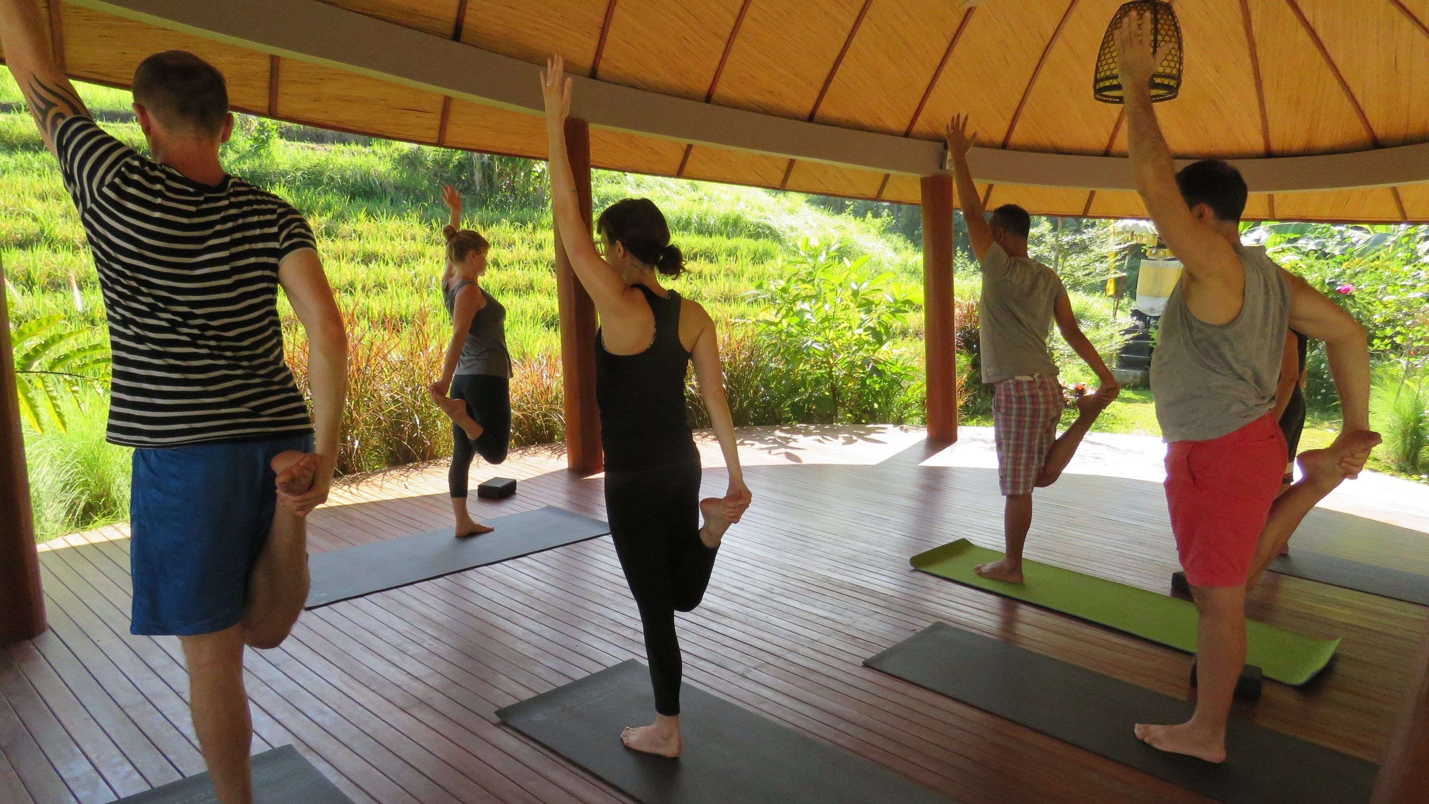 Un groupe de personnes pratiquant le yoga sur des tapis en bois, sous une structure en bois abritée, avec de la verdure visible à l’extérieur.