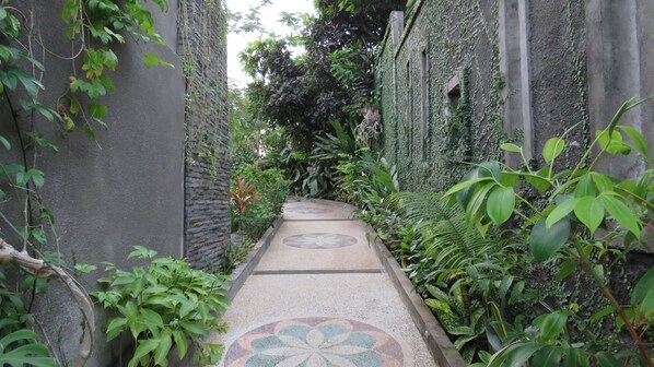 A narrow pathway with patterned tiles leading through a lush garden area, surrounded by dense foliage and stone walls.