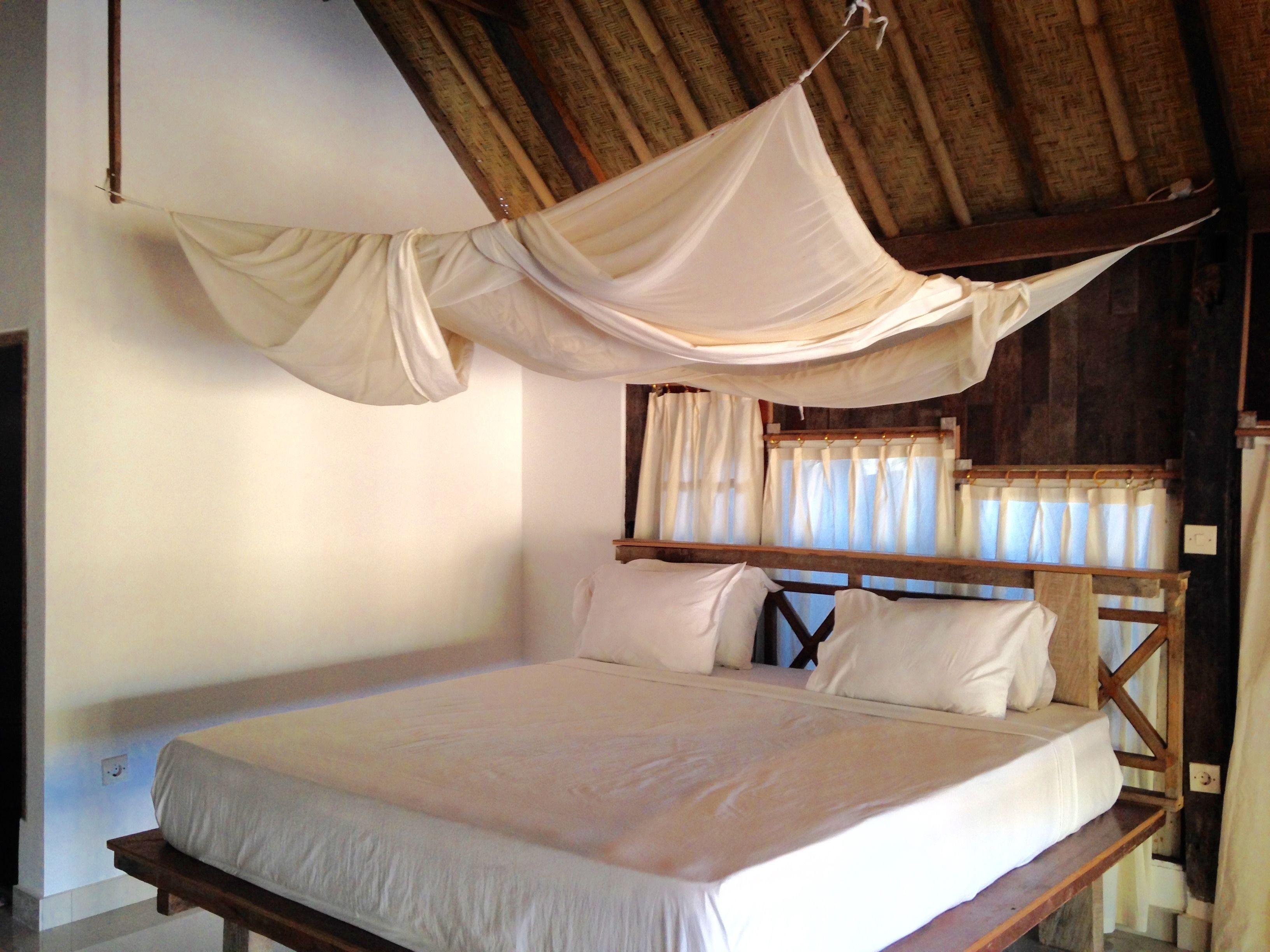 A bed with a wooden frame and white bedding, a wooden headboard with curtains, and a thatched roof.