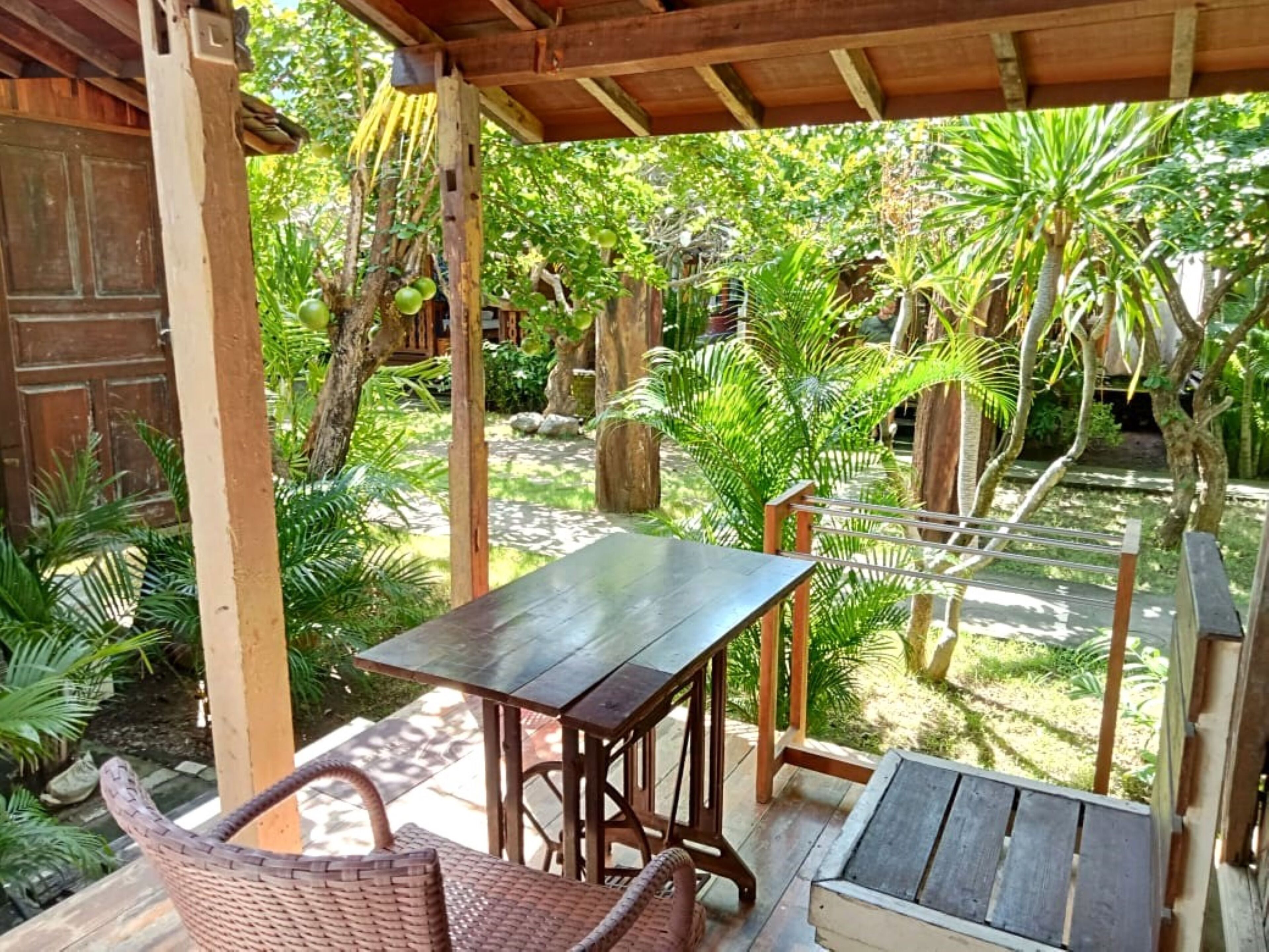 A covered outdoor seating area with a table and chairs, surrounded by lush greenery and trees.
