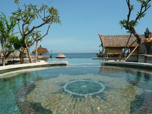 A clear infinity pool with a view of the ocean, surrounded by tropical trees and thatched-roof structures. - Classic Beach Villas (Karangasem)