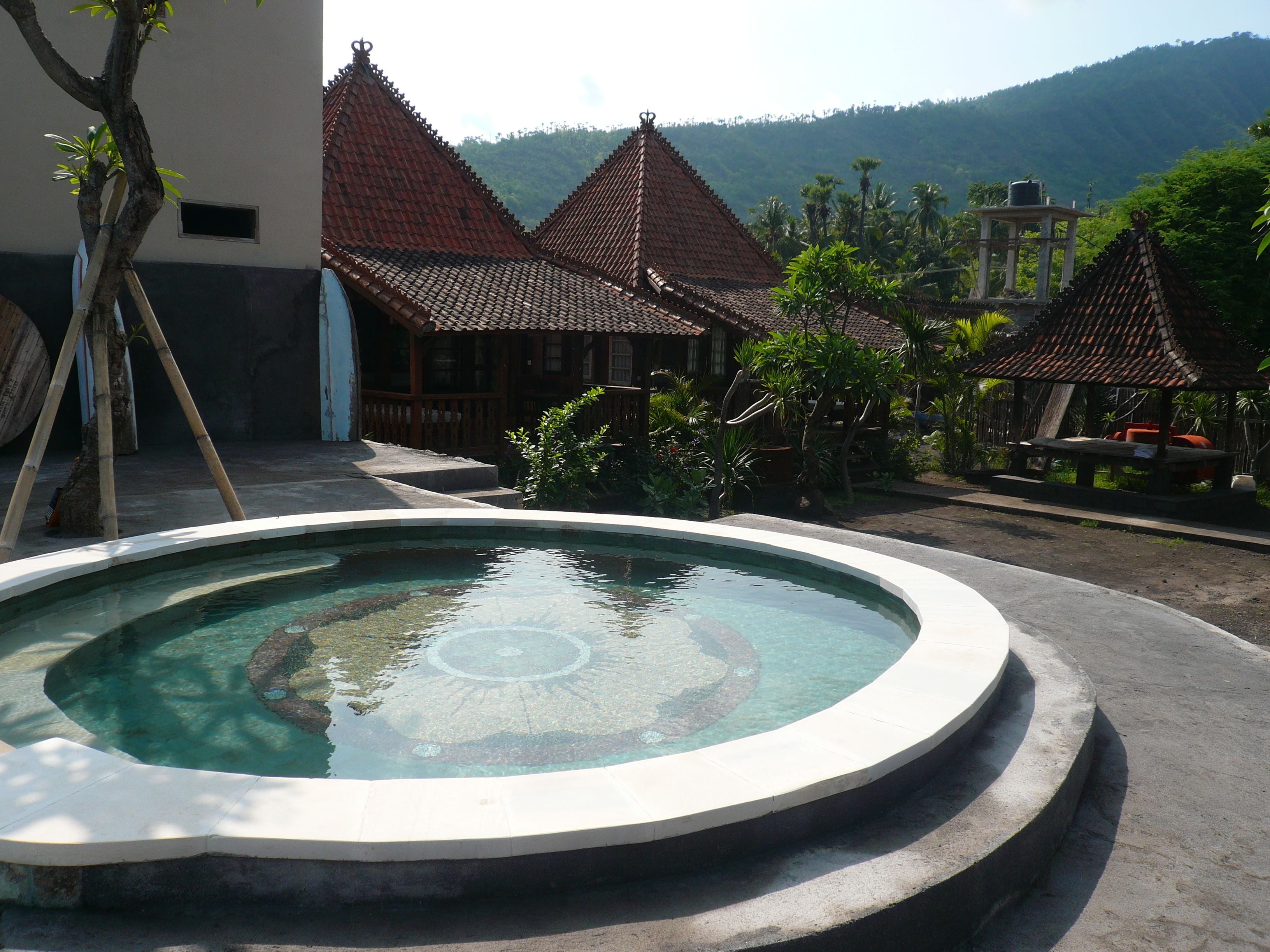 A swimming pool with a circular design, surrounded by a concrete deck. There are traditional buildings with red tiled roofs in the background.