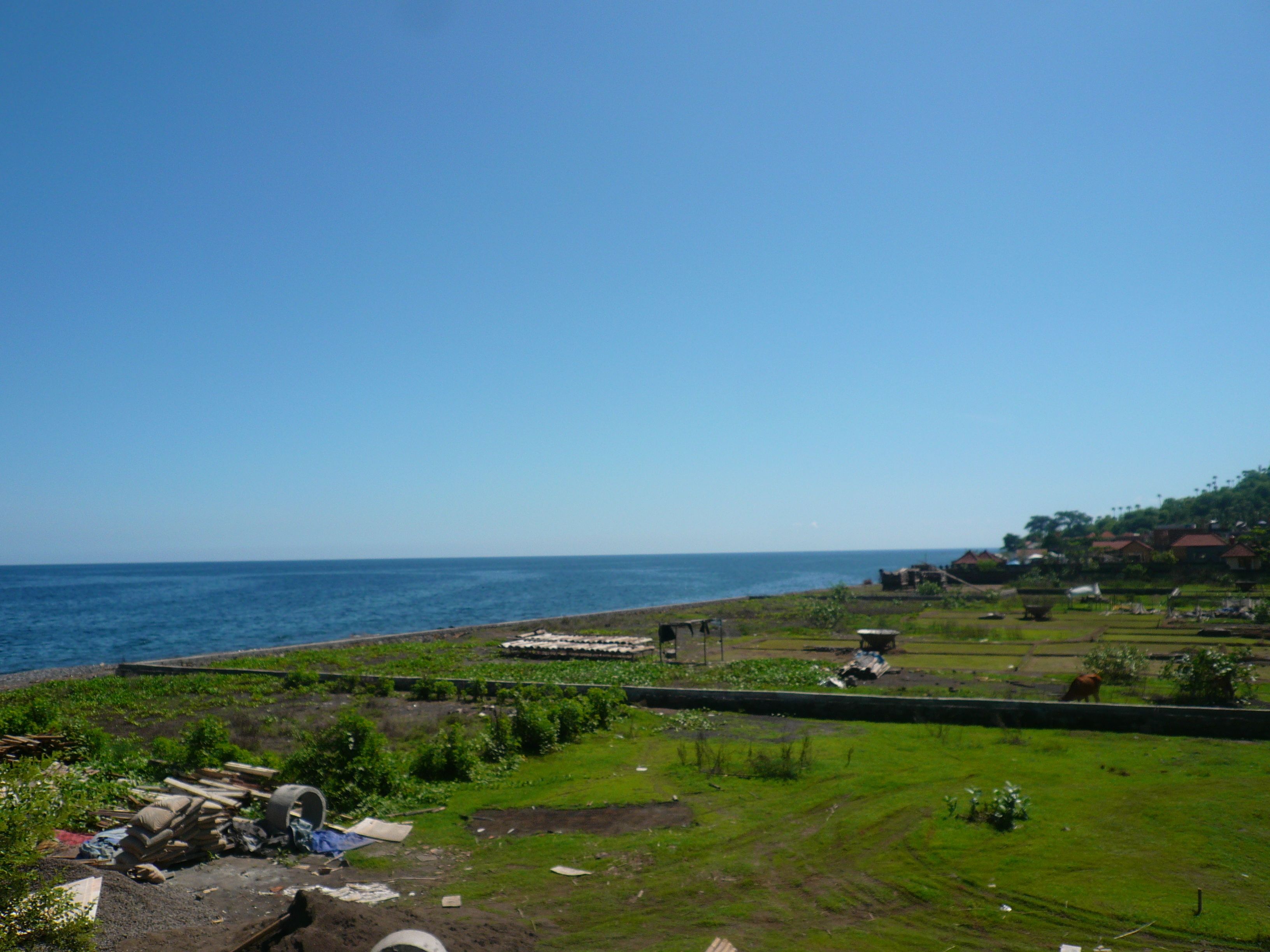 Un paisaje costero con vista despejada al mar, una zona cubierta de césped con algunos escombros, y un grupo de edificios a lo lejos.