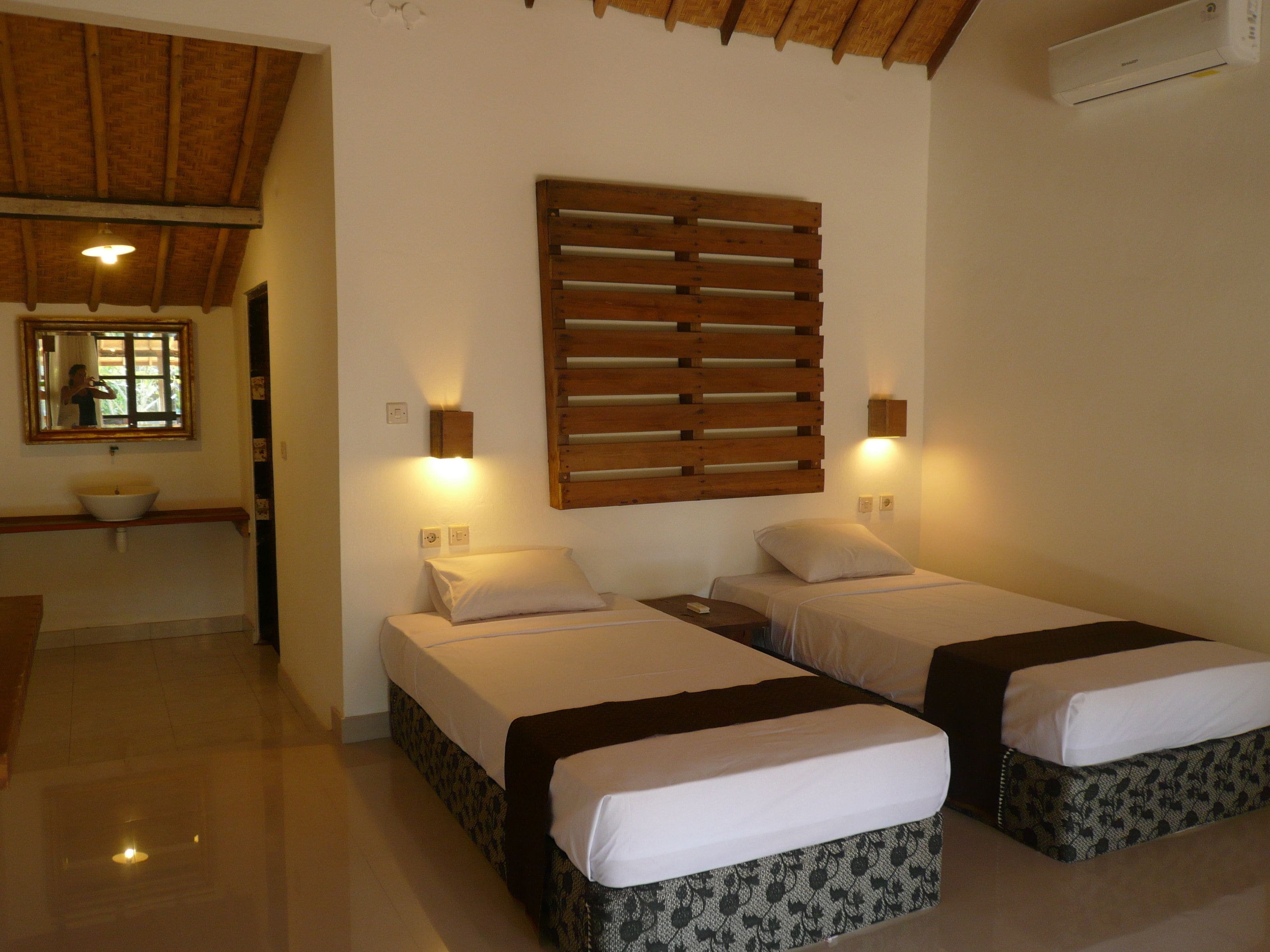 Habitación de hotel con dos camas, cabecero de madera con listones, un espejo grande, un lavamanos y una ventana.