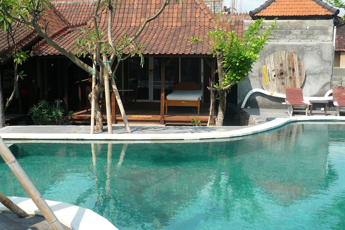 A clear swimming pool with a wooden deck and lounge chairs, surrounded by a traditional building with a tiled roof.