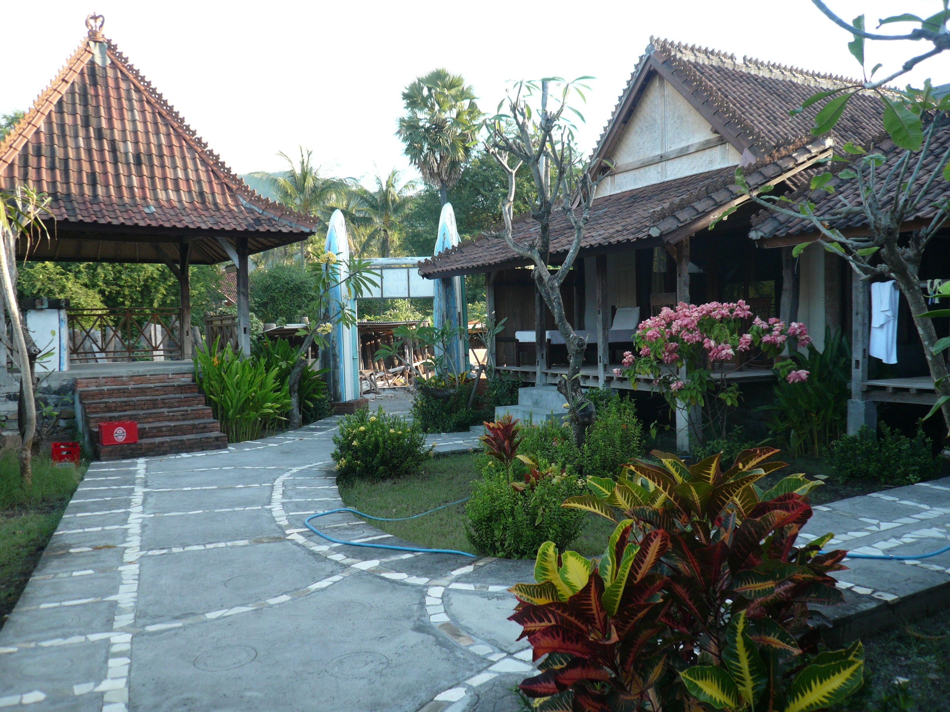 A traditional wooden structure with a tiled roof, surrounded by lush greenery and a stone pathway leading to a covered entrance.