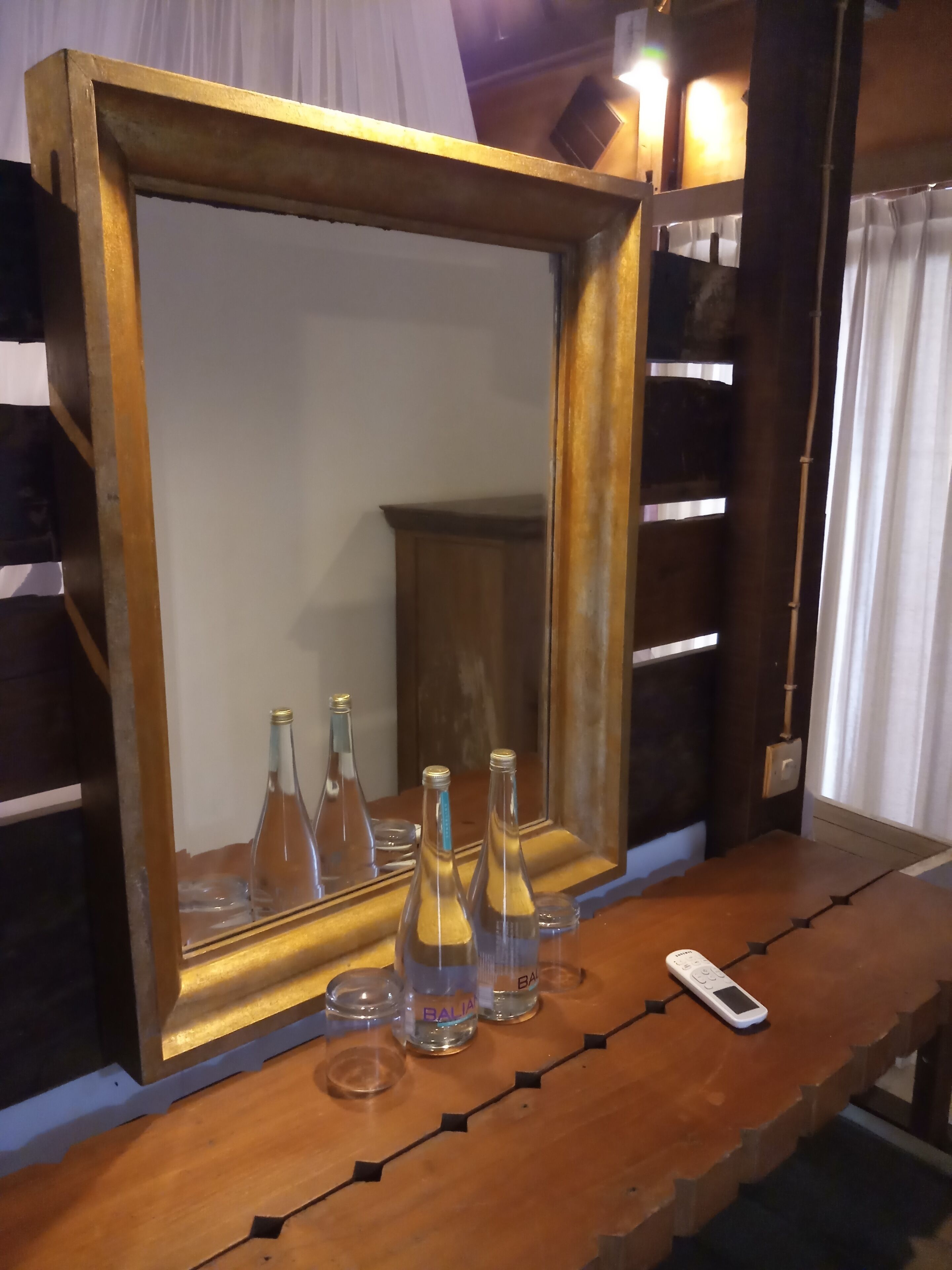 Un espejo con marco de madera sobre una mesa de madera, tres botellas transparentes con etiquetas y un control remoto.