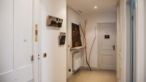 A hallway with white walls, a radiator, and a door with a 'WC' sign. There are artworks on the wall and a branch with lights. - Félix dOrt (Lyon)