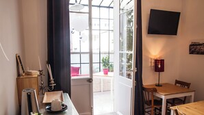 A room with a wooden table, a chair, a small table with a cup and saucer, a potted plant, a wall-mounted TV, and a door leading to an outdoor area. - Félix dOrt (Lyon)