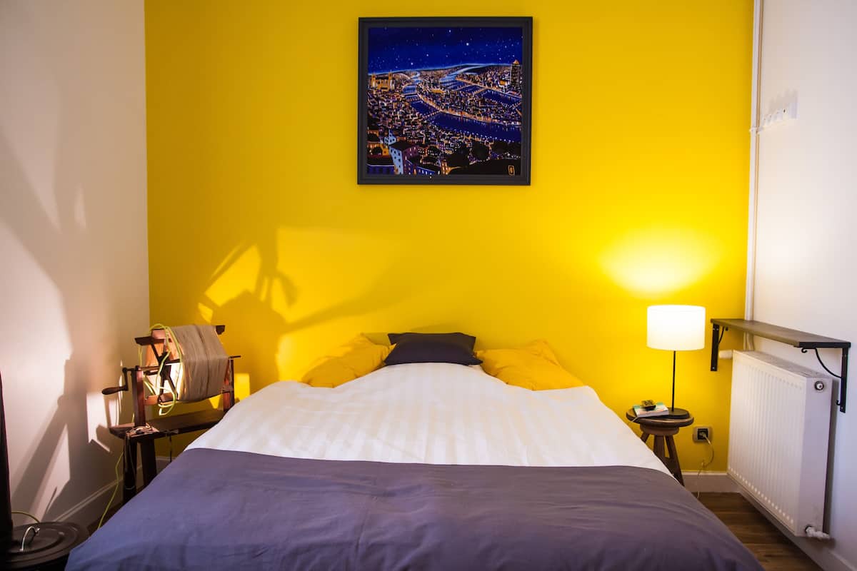 Une chambre d’hôtel avec un lit, une affiche représentant un paysage urbain nocturne, une lampe et une petite table avec une autre lampe.