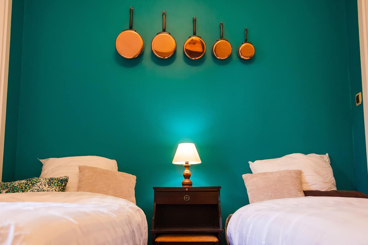 Une chambre avec des murs de couleur turquoise, deux lits avec une literie blanche, une table de chevet avec une lampe et un présentoir mural avec cinq objets ronds.