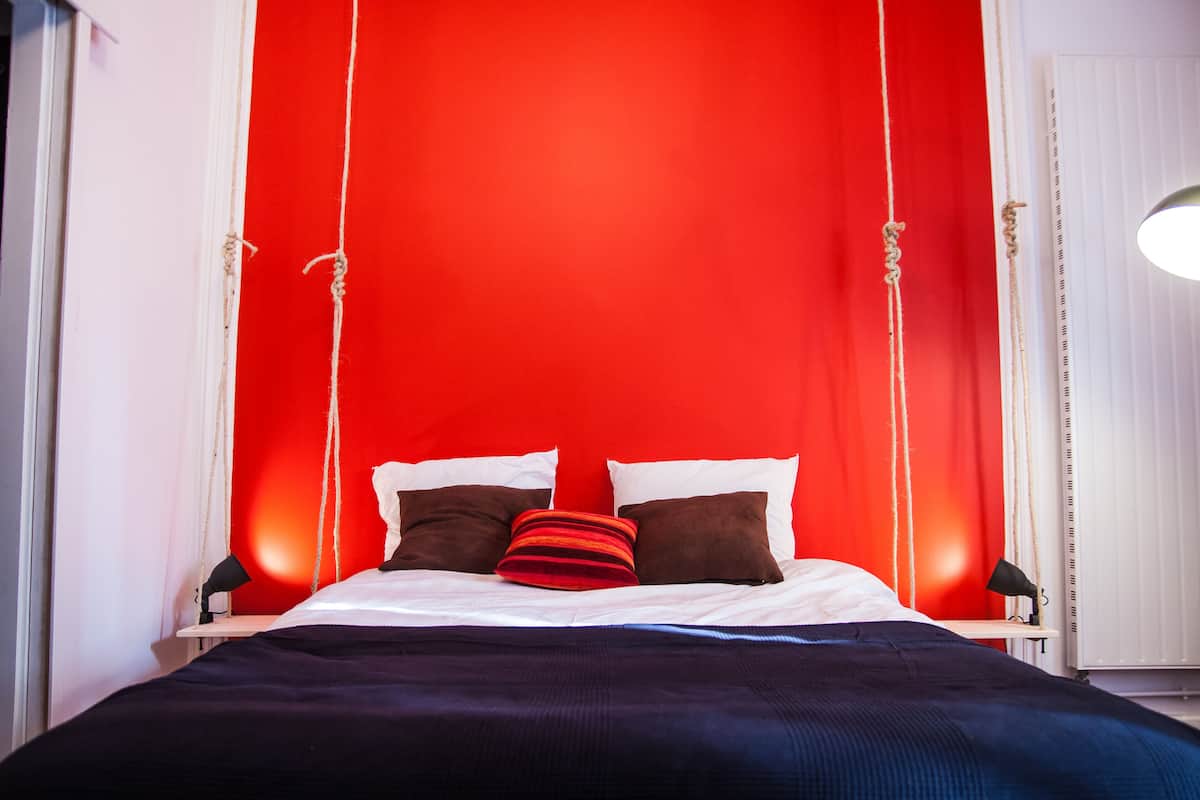 Un lit avec une literie blanche et marron, un coussin rouge qui apporte une touche de couleur, et une lampe noire posée sur une table de chevet.