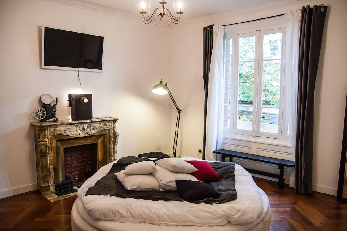 Une chambre à coucher avec un lit, une cheminée, un téléviseur et une fenêtre avec des rideaux.