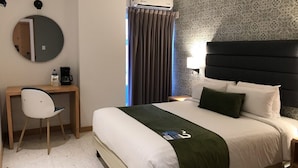 Una habitación de hotel con cama, escritorio, silla, espejo redondo y unidad de aire acondicionado.