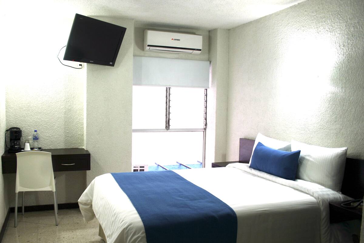 Una habitación de hotel con cama, escritorio, silla, televisión y aire acondicionado.