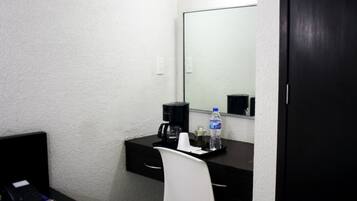 Una habitación pequeña y privada con escritorio, silla y un espejo grande. Hay una cafetera, una botella de agua y un teléfono sobre el escritorio.