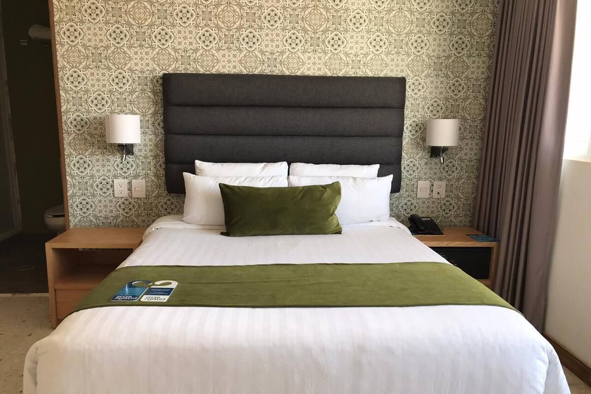 Habitación de hotel con una cama grande, una manta verde y cabecera. Hay dos mesitas de noche con lámparas y teléfono.