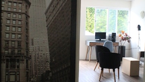Una habitación con un cuadro grande de un paisaje urbano, un escritorio con una computadora, una silla y un florero con flores.