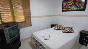Habitación de hotel con cama, televisor, cuadro y ventana con persianas.