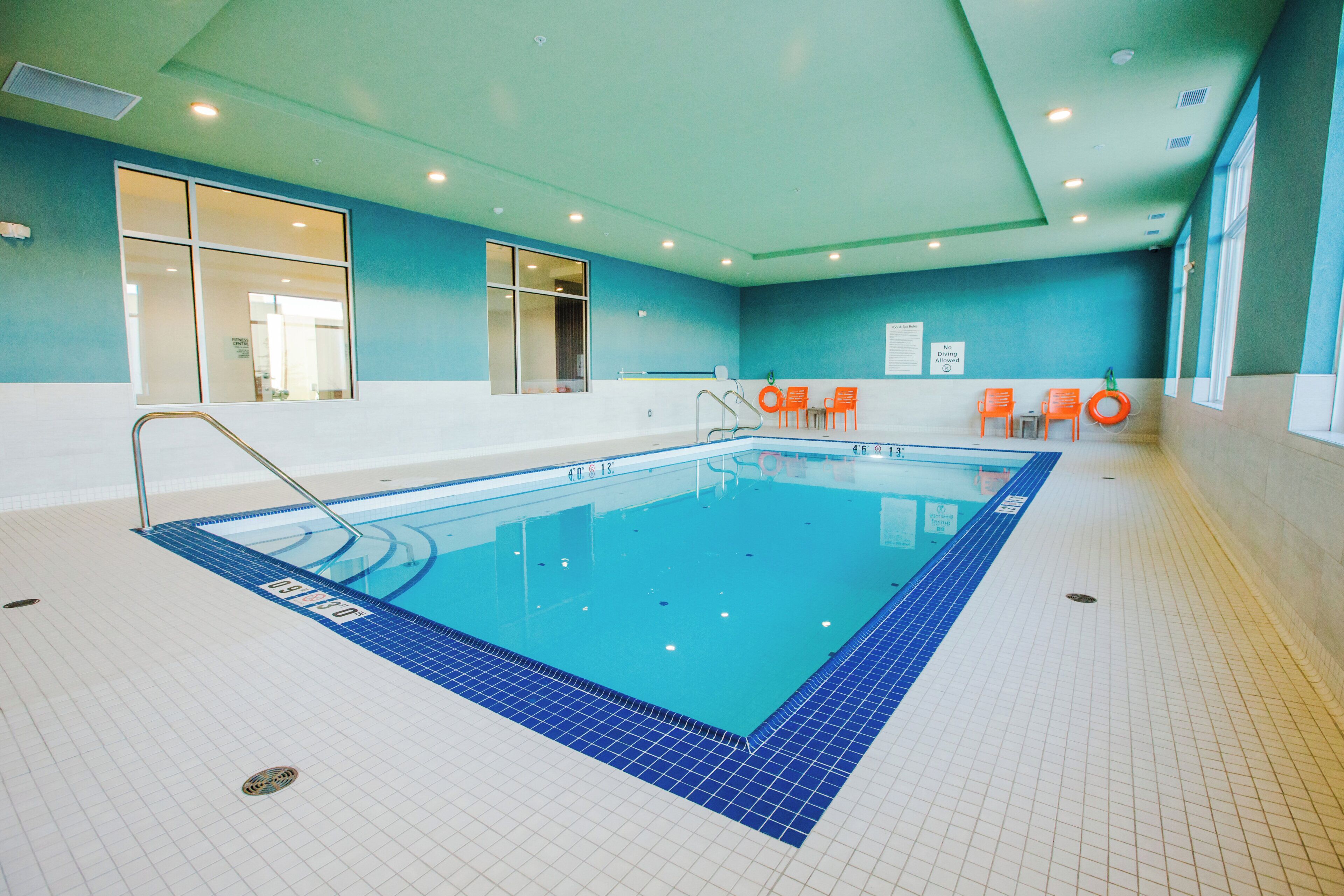 An indoor swimming pool with a tiled floor, a diving board, and orange chairs along the wall.