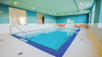 An indoor swimming pool with a tiled floor, a diving board, and orange chairs along the wall.