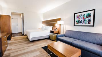 Une chambre d’hôtel moderne comprenant un lit, un canapé, une table basse en bois, une table de chevet et un tableau abstrait encadré accroché au mur.