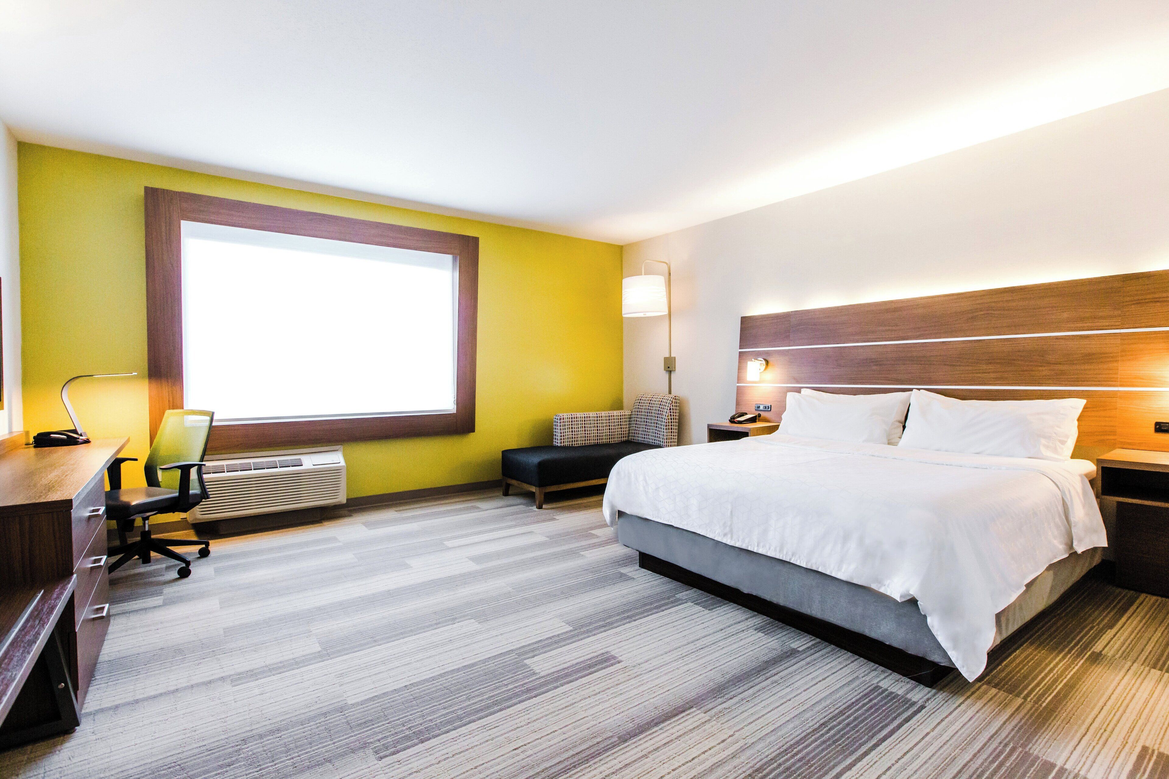 A hotel room with a large bed, a desk with a chair, a window, and a wall-mounted lamp.