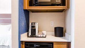 Una kitchenette compacta con microondas, cafetera y una taza negra sobre una encimera blanca.