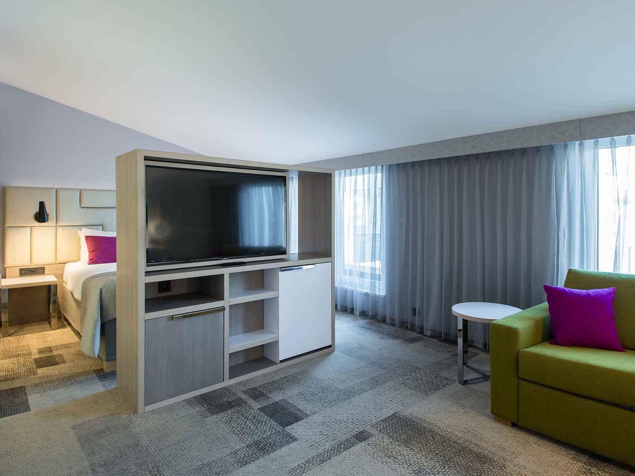 Habitación de hotel con cama, televisor, un refrigerador pequeño, sofá y ventana con cortinas.