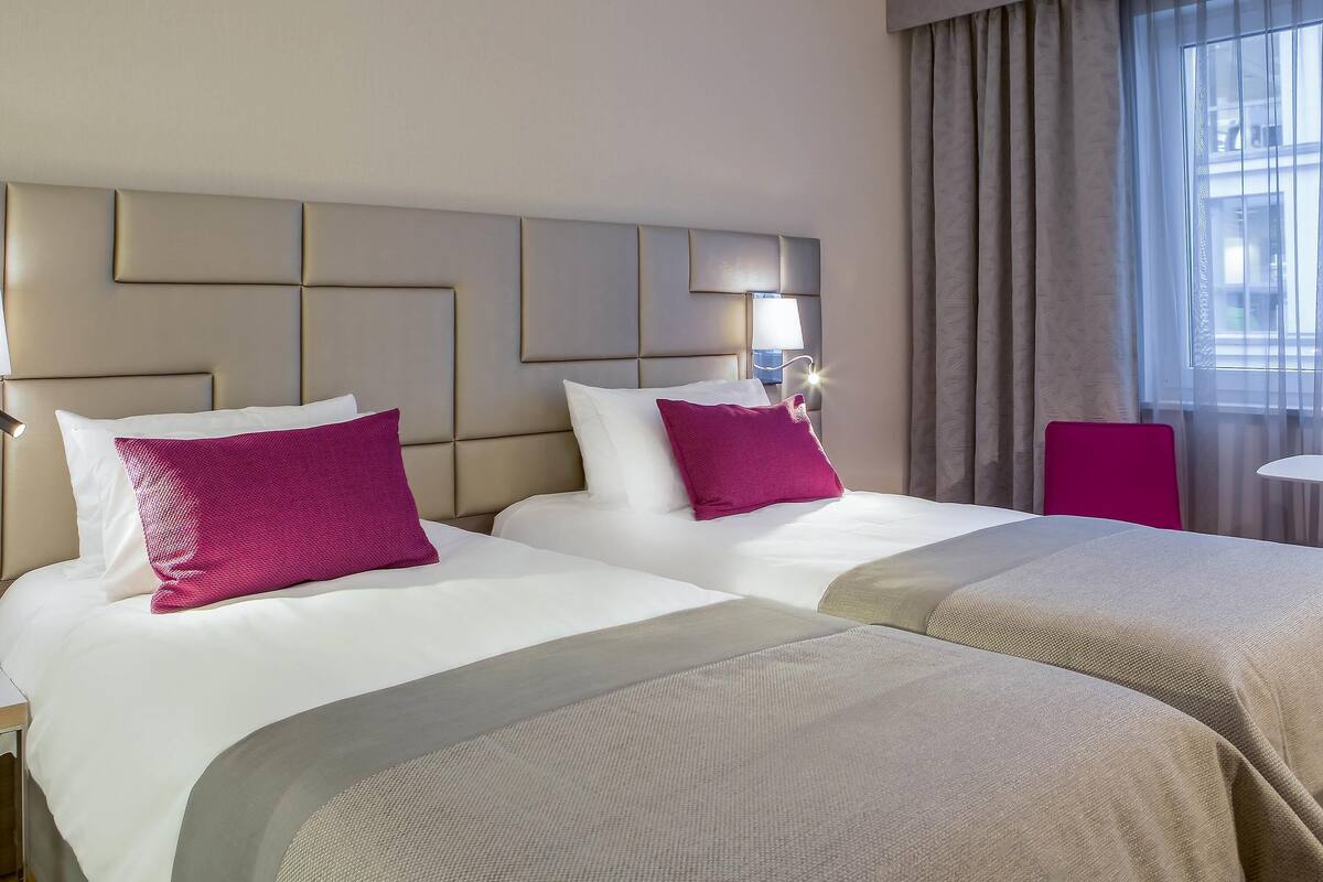 Une chambre d’hôtel avec deux lits, une tête de lit, une table de chevet, une lampe, une chaise et une fenêtre avec des rideaux.