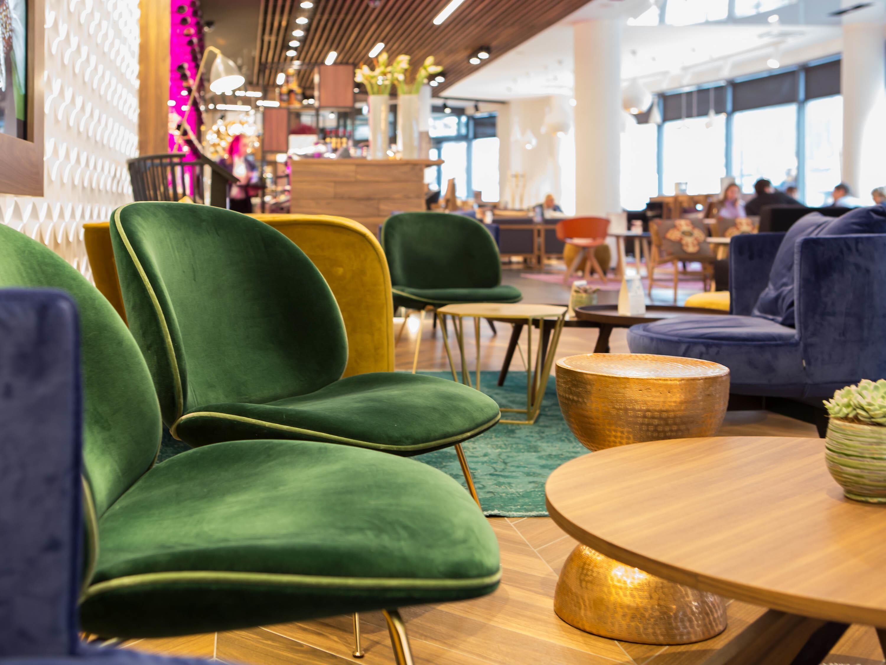 Un espace de détente moderne avec des fauteuils verts et jaunes, des tables en bois et une grande fenêtre donnant sur l’intérieur.