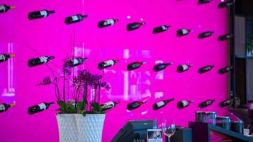 A wall with numerous wine bottles arranged in a pattern, a potted plant, and a few wine glasses on a counter.