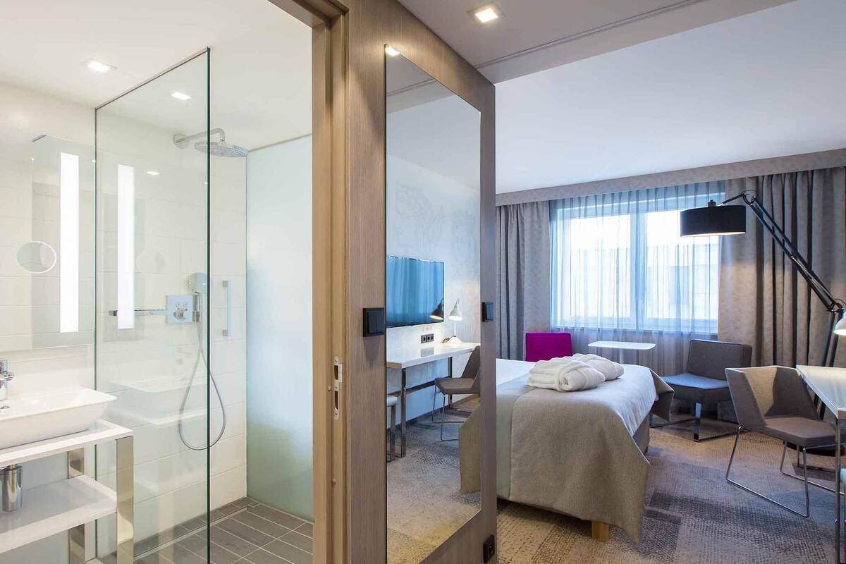 Une chambre d’hôtel moderne dotée d’une douche en verre, d’un bureau avec une chaise, d’un lit avec une couverture et d’un grand miroir.