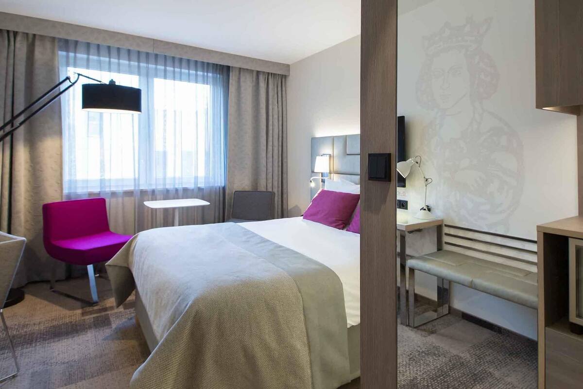 Een hotelkamer met een groot bed, een bureau, een stoel, een lamp en een raam met gordijnen.