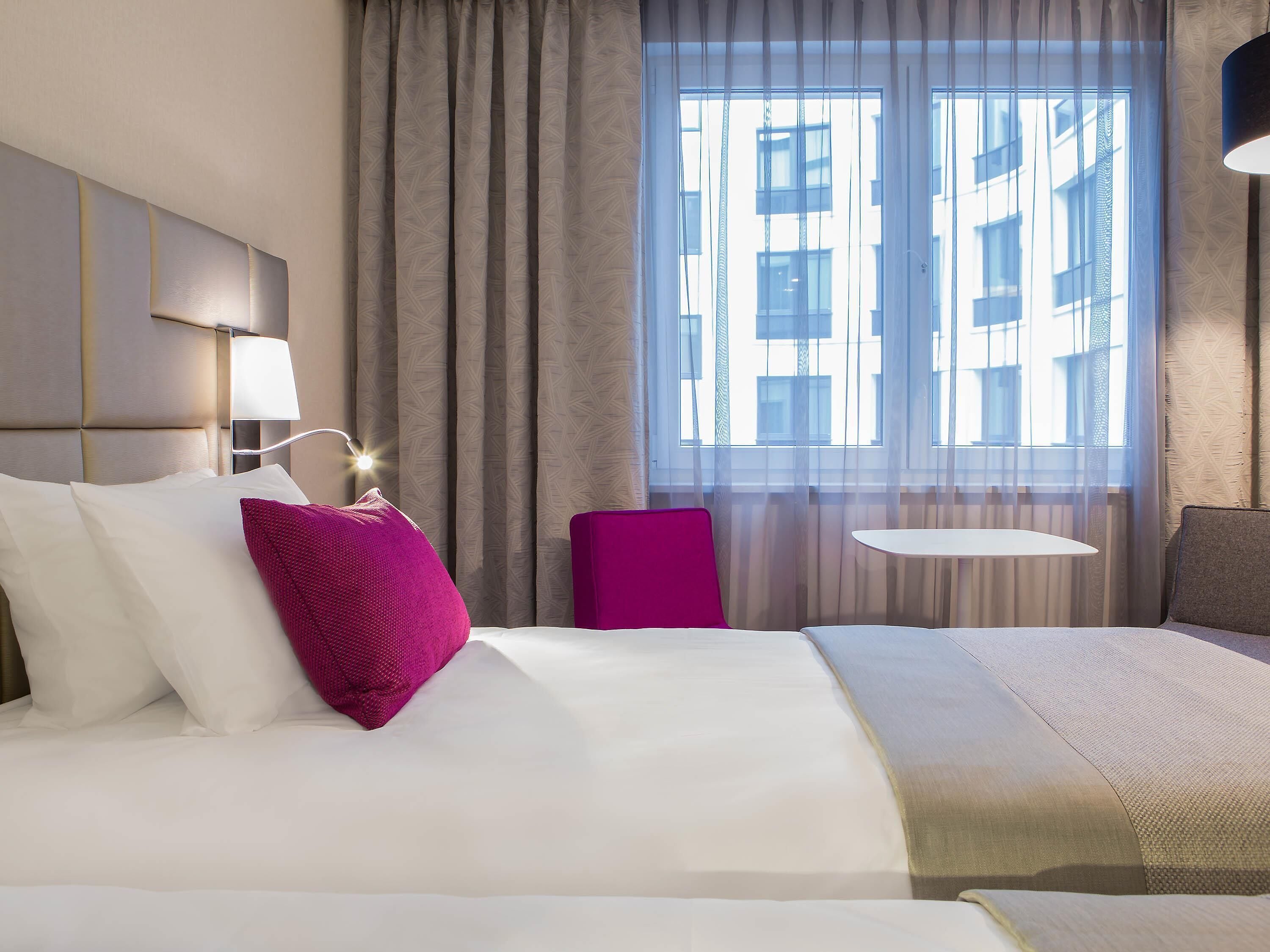 Une chambre d’hôtel avec un lit, une lampe de chevet, une fenêtre avec des rideaux et une vue sur les bâtiments environnants.