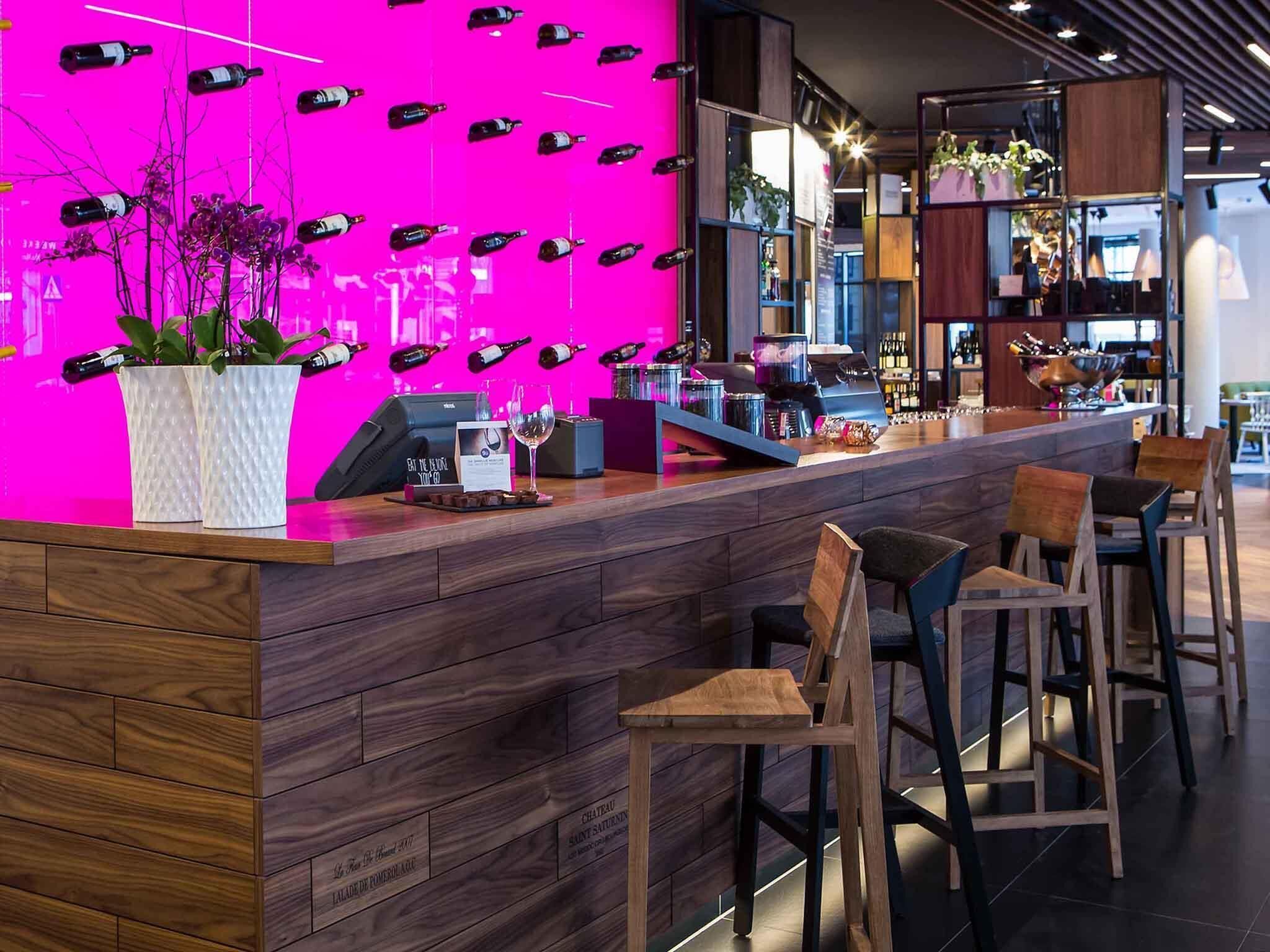 A bar area with a wooden counter, bar stools, and a vibrant pink wall with hanging bottles.